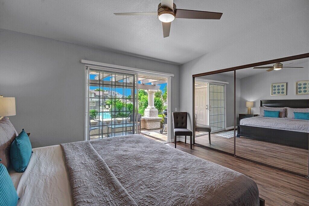 A bedroom with a large bed, a ceiling fan, a view of the outdoors, and a sliding glass door leading to a patio.