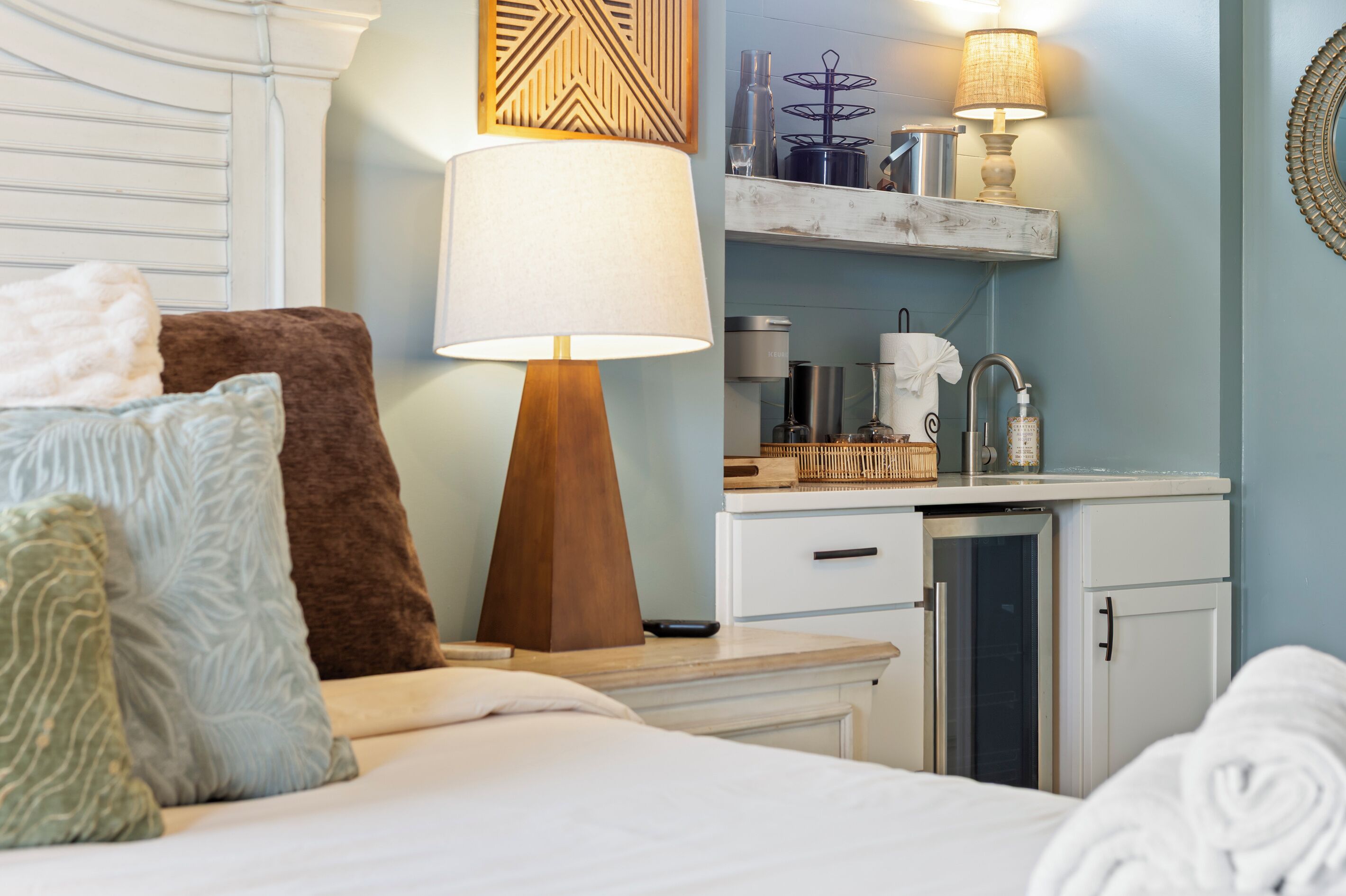 A hotel room with a bed, pillows, a bedside table, a lamp, a small kitchenette, and a mirror.