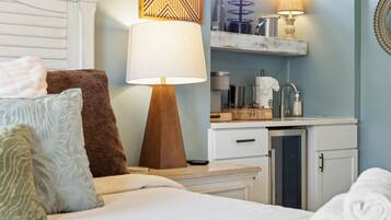 A hotel room with a bed, pillows, a bedside table, a lamp, a small kitchenette, and a mirror.