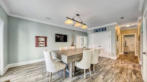 A dining area with a wooden table and chairs, a television mounted on the wall, and a hallway leading to a door labeled 'HOME'. - Walk to Beach: 9BR Miramar Retreat - Pool and Grill (Destin)