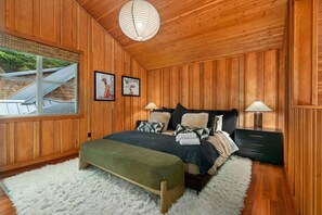 Een slaapkamer met een houten lambrisering, een bed met veel kussens, een nachtkastje met een lamp en een raam met uitzicht op een dak.