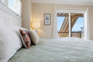 A bedroom with a bed, a bedside lamp, a framed picture, and a view of a balcony with wooden railings. - Oceanfront | Private Boardwalk | Gazebo (North Topsail Beach)