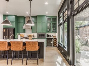 A modern kitchen with green cabinets, a central island with bar stools, pendant lighting, and a large window offering a view of the outdoors. - Chic Crown Heights Retreat W/ Fire Pit, Near Paseo (Oklahoma City)