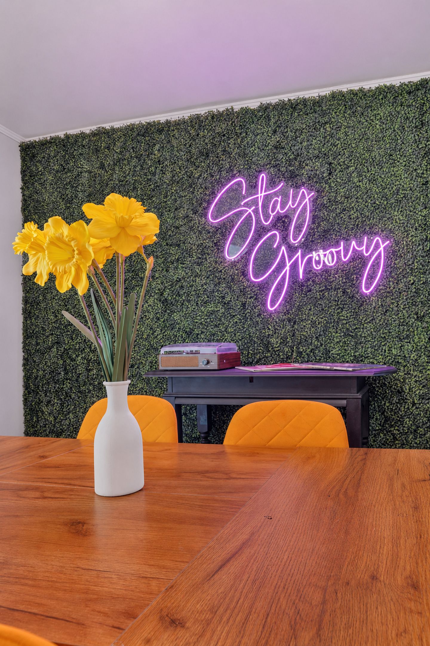 Une pièce avec une table en bois, un vase blanc orné de fleurs jaunes, un panneau lumineux au néon et un mur recouvert de feuillage vert.