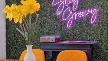 Ein Zimmer mit einem Holztisch, einer weißen Vase mit gelben Blumen, einem Neonlicht und einer Wand, die mit Grünpflanzen bedeckt ist.
