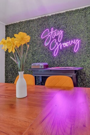 A room with a neon sign that reads 'Stay Groovy,' a wooden table, a white vase with flowers, and a wall covered in foliage. - The Peach Haus ~ Groovy Stay With Screened Porch (Tifton)