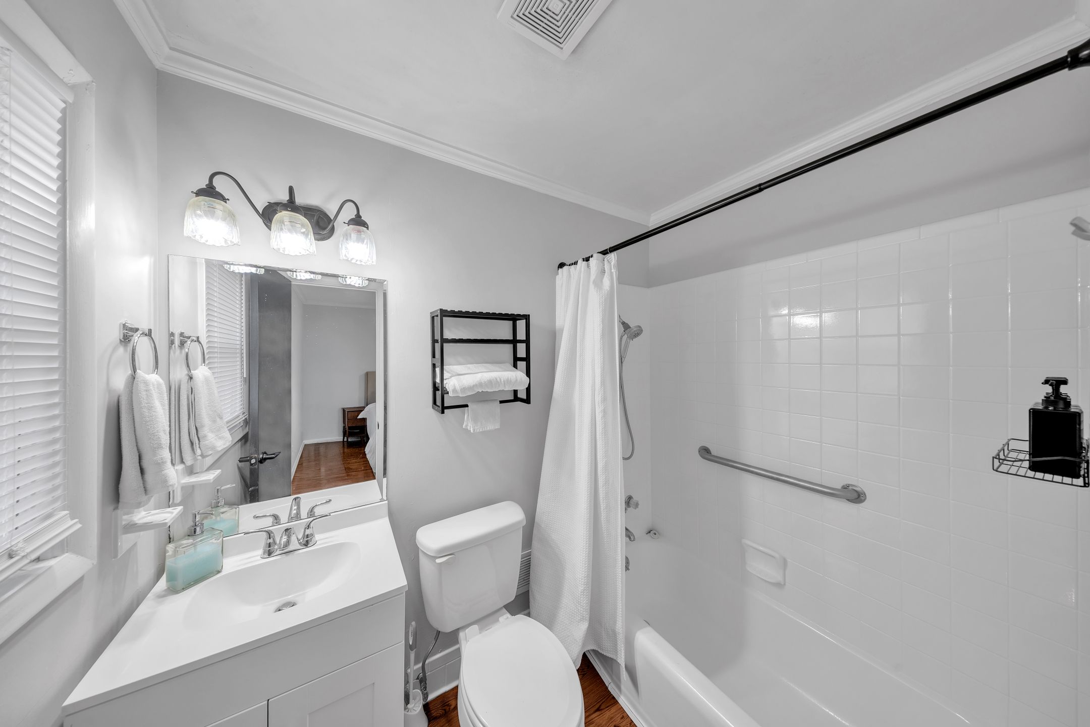 Un baño con lavamanos blanco, espejo y retrete. Cuenta con una ducha con puerta de vidrio y una barandilla.