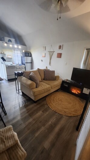 Een woonkamer met een bank, een flatscreen televisie en een open haard. In de achtergrond is een keuken met een magnetron en een koelkast.