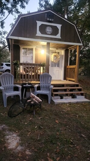 Een klein houten huisje met een veranda, twee Adirondack-stoelen, een vuurkorf en een welkomstbord.