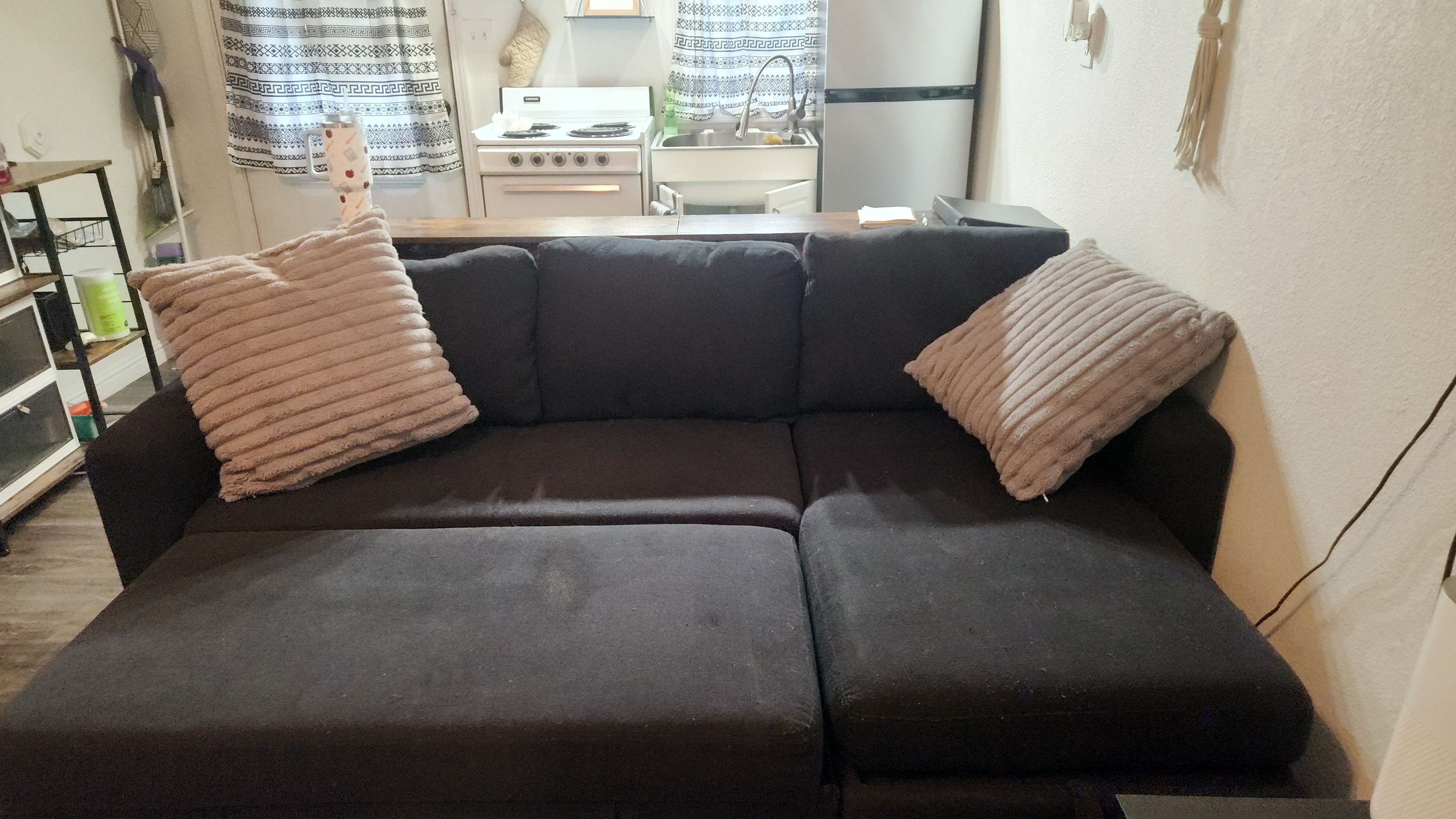 Um sofá escuro com duas almofadas, uma área de cozinha ao fundo e uma cortina estampada.