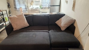 A dark-colored sofa with two pillows, a kitchen area in the background, and a patterned curtain.