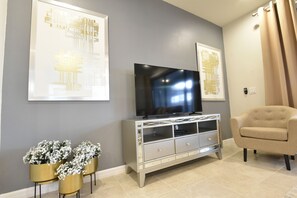 Un salón con una televisión de pantalla plana sobre un mueble plateado, un sillón color crema y tres jarrones dorados con flores blancas en el suelo.