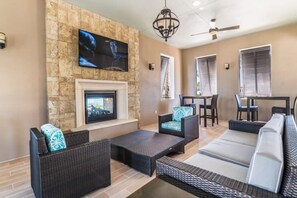 A modern living room with a stone wall, a fireplace, and a television mounted above it. There are wicker sofas and a dining area with a table and chairs. - Princess/StarWars 5-bedroom house in Orlando near Parks (Kissimmee)