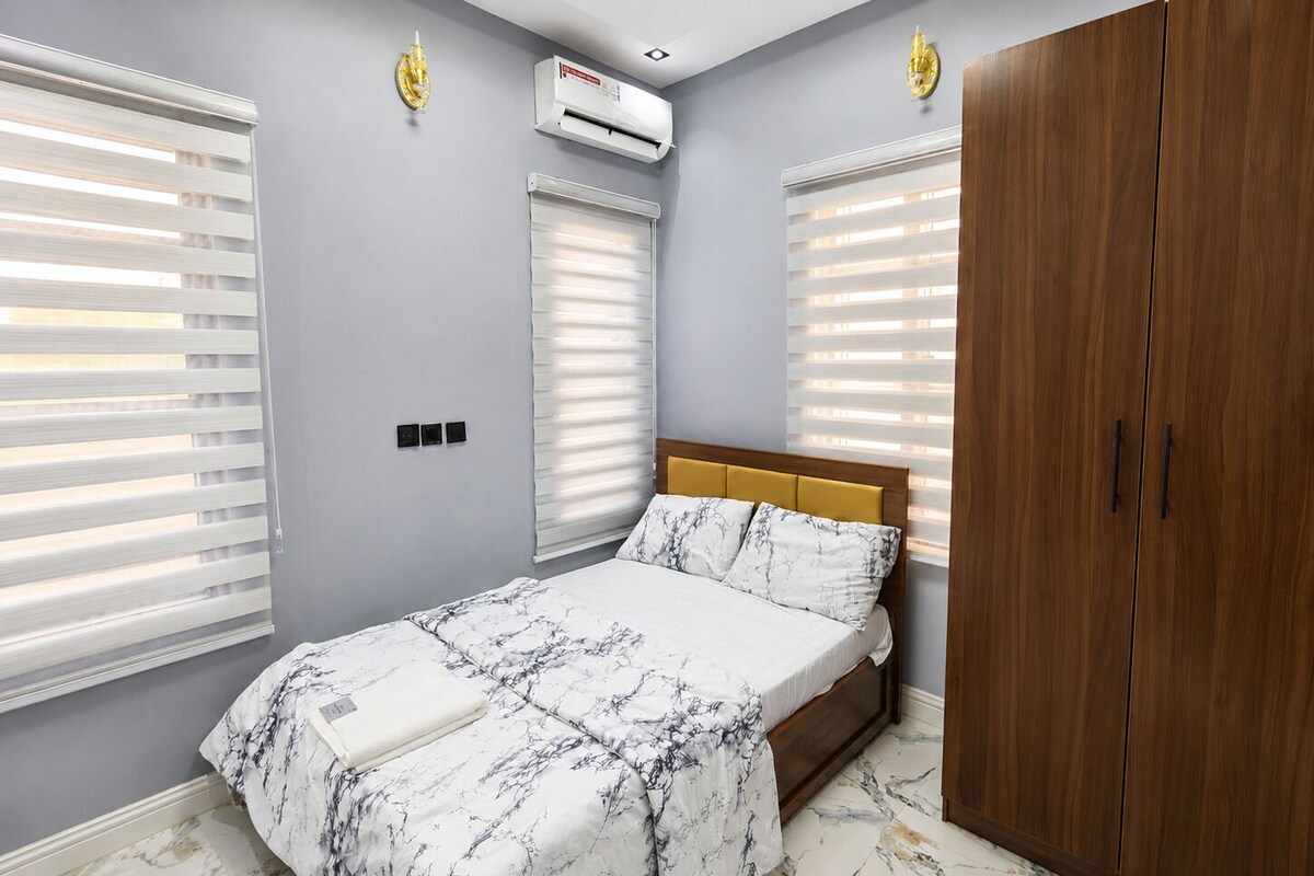 Ein Schlafzimmer mit einem Bett, zwei Fenstern mit Jalousien, einer Klimaanlage und einem Kleiderschrank.