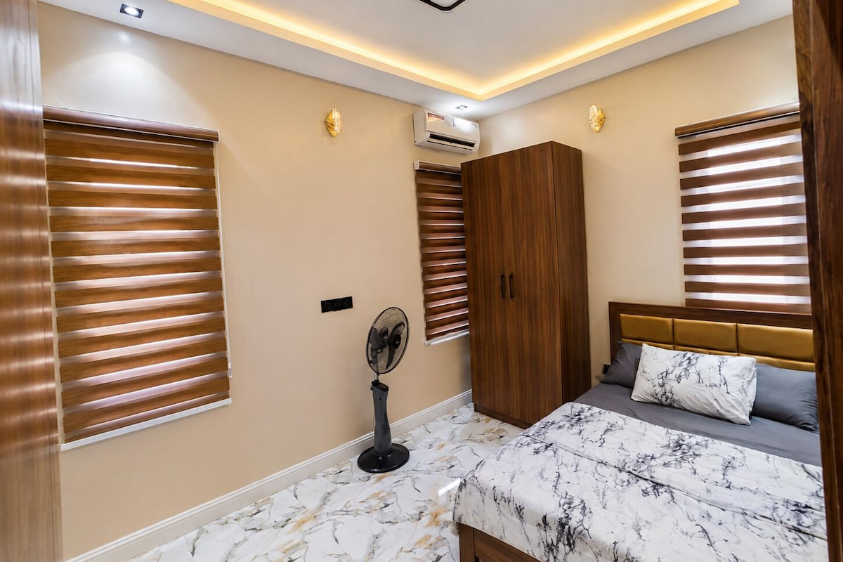Ein Schlafzimmer mit einem Bett, einem Kleiderschrank, einem Standventilator und einer Deckenleuchte.