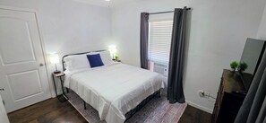 A bedroom with a bed, bedside tables, a lamp, a window with curtains, a radiator, and a TV on a stand.