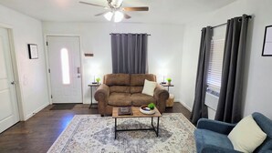 A living room with a brown sofa, a blue armchair, a coffee table, a rug, a ceiling fan, and a window with curtains.