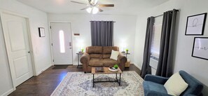 A living room with a brown sofa, a blue armchair, a coffee table, a rug, a ceiling fan, and a window with curtains.
