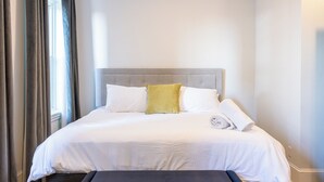 Un letto rifatto con un poggiatesta grigio, cuscini bianchi e gialli, e un asciugamano piegato.