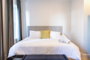 Una cama bien hecha con cabecera gris, almohadas blancas y amarillas, y una toalla doblada.