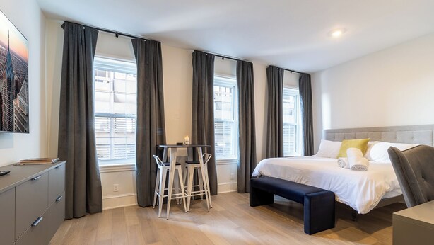 Una camera d'hotel con un letto, un comodino, una sedia, una scrivania e vista sulla città attraverso ampie finestre.