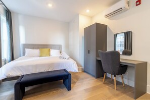 Habitación de hotel con cama, mesita de noche, banco, escritorio con silla y unidad de aire acondicionado.