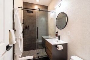 Un baño moderno con ducha envuelta en vidrio, un espejo redondo, un lavamanos blanco y un inodoro.