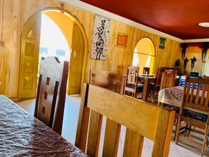 Una habitación con paredes y puertas de madera, una cama con colcha estampada, una zona de comedor con sillas y una mesa, y un cuadro en la pared.
