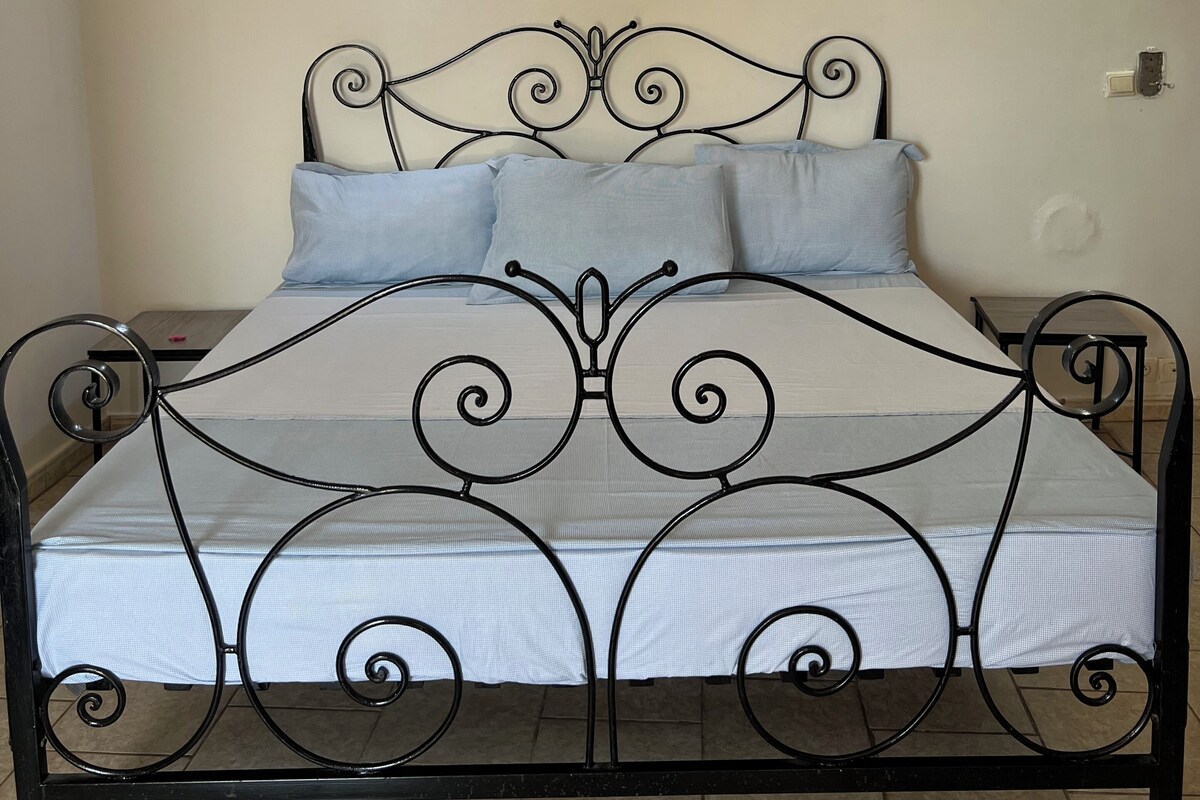 Una cama con estructura metálica y colchón blanco, dos lámparas fijadas a la pared y un equipo de aire acondicionado en la pared.