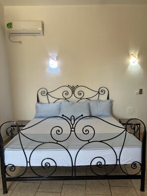 Una cama con estructura metálica y colchón blanco, dos lámparas fijadas a la pared y un equipo de aire acondicionado en la pared.