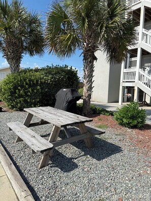 A wooden picnic table with benches, surrounded by gravel and small plants, with palm trees and a building in the background. - NEW! Surf City 2BR Beach Escape Walk Everywhere Labor Day Available (Surf City)