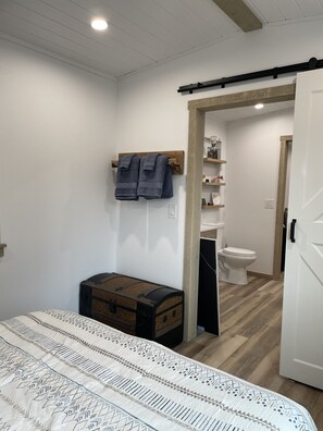 A bedroom with a bed, a chest of drawers, a wooden bench, a towel rack, and a doorway leading to a bathroom with a toilet and a sink. - Railroad Retreat in Moose Pass – Car 0037 (Moose Pass)