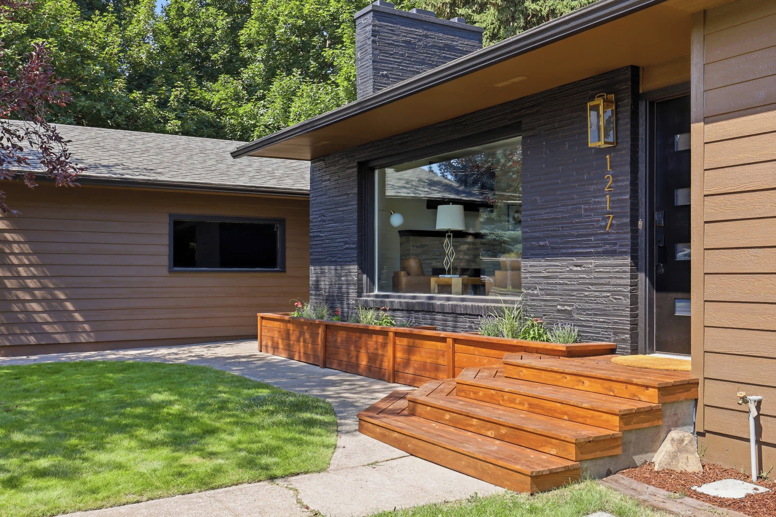 A modern house with a stone facade, a large window, and a wooden deck with steps leading to the entrance.