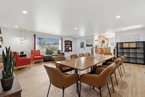 A modern dining room with a long table and multiple chairs, a kitchen area with wooden cabinets, and a large window with a view of greenery.
