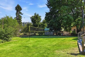 Área gramada bem conservada, com uma rede de voleibol, cercada por árvores e um edifício ao fundo.