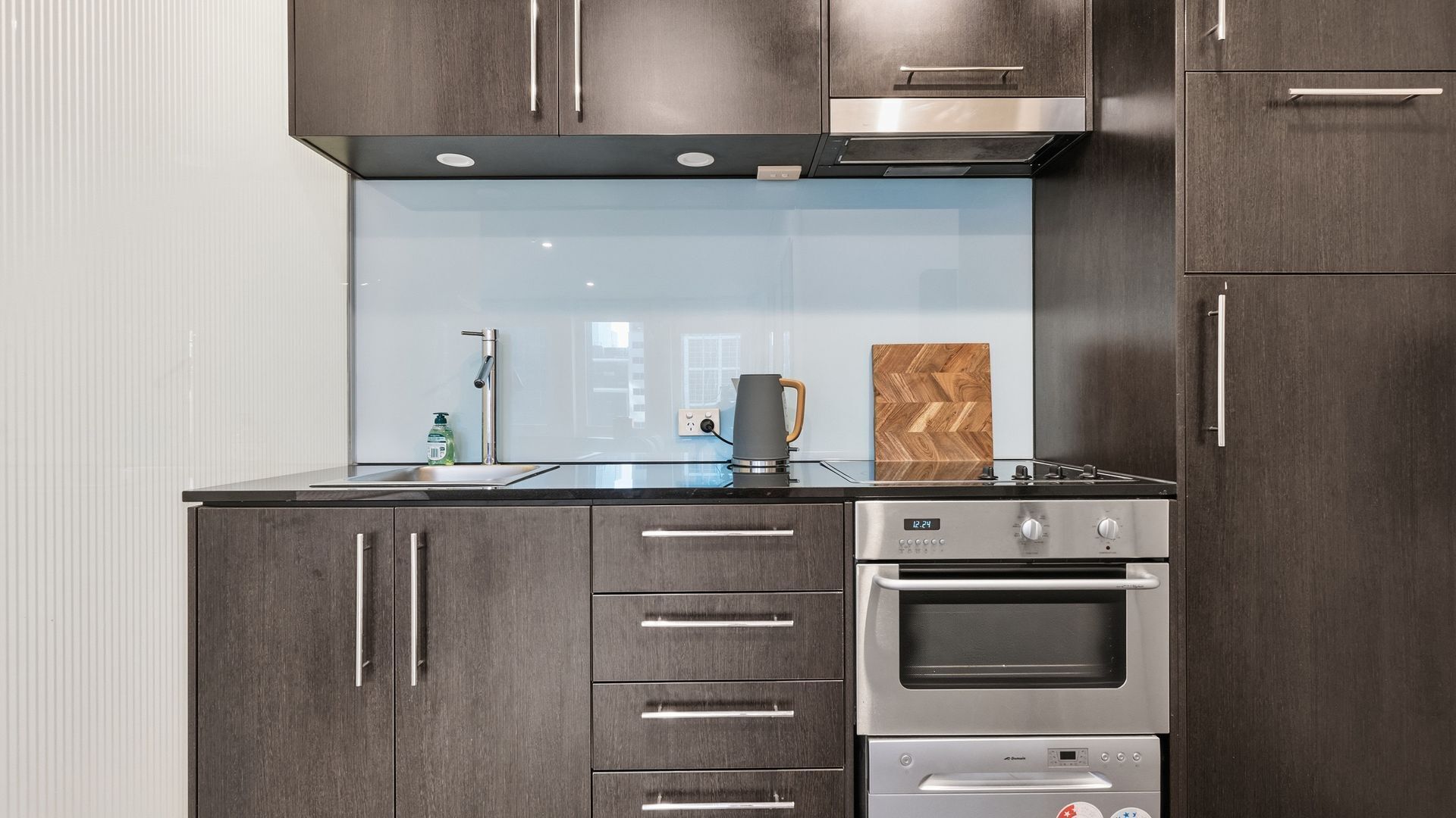 Een moderne keuken met donkere houtkasten, roestvrijstalen apparatuur, een glazen spatwand en een inbouwoven.