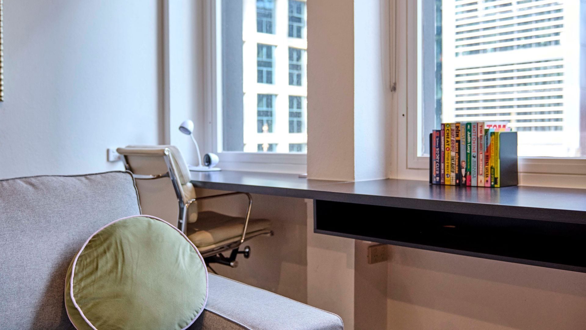 Un espace de bureau moderne comprenant un bureau, une chaise, un canapé avec un coussin et une fenêtre avec des stores.