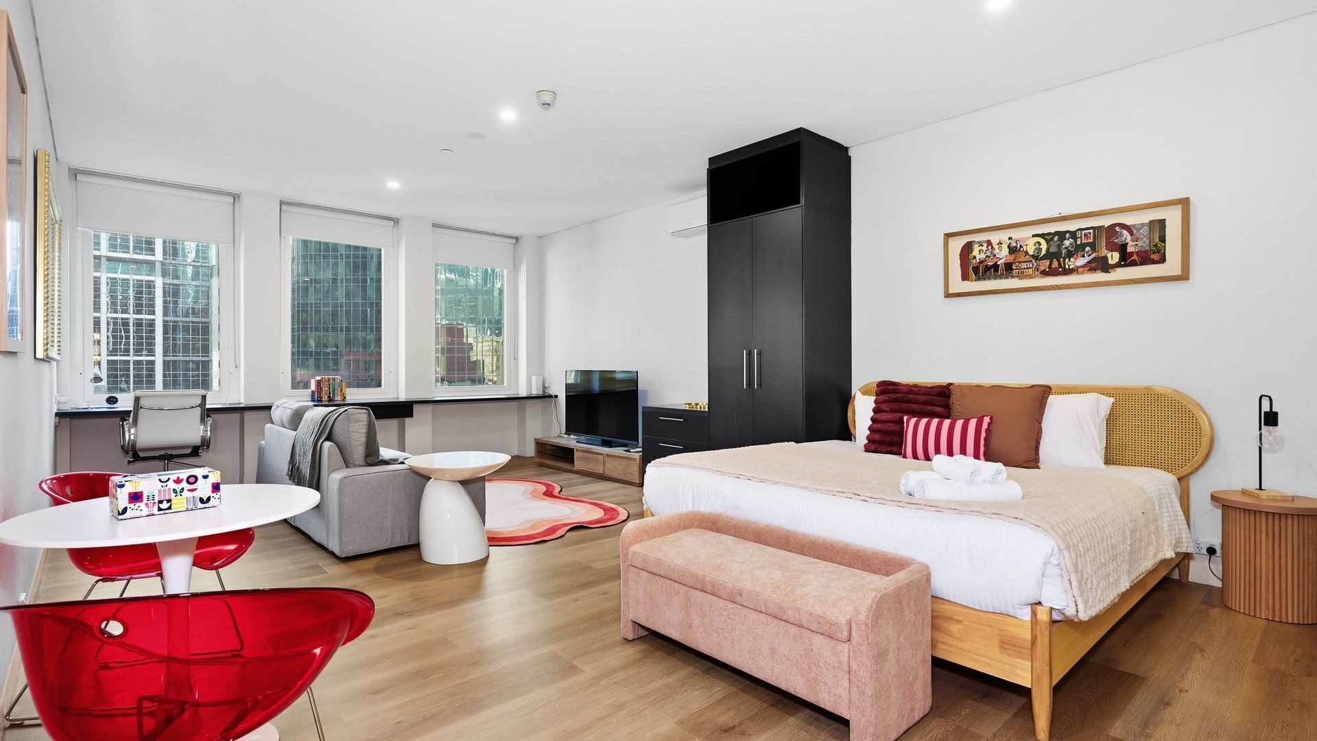A modern hotel room with a bed, a television, a sofa, a red chair, and a painting on the wall.