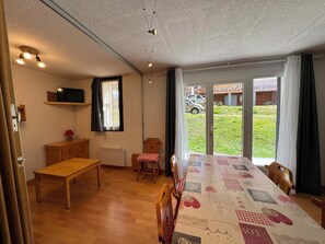 A room with a wooden table, a chair, a television on a shelf, and a view of the outside through a glass door. - Appartement 8 pers - 3 chambres - Résidence le Christiana - Centre village (Risoul 1850)