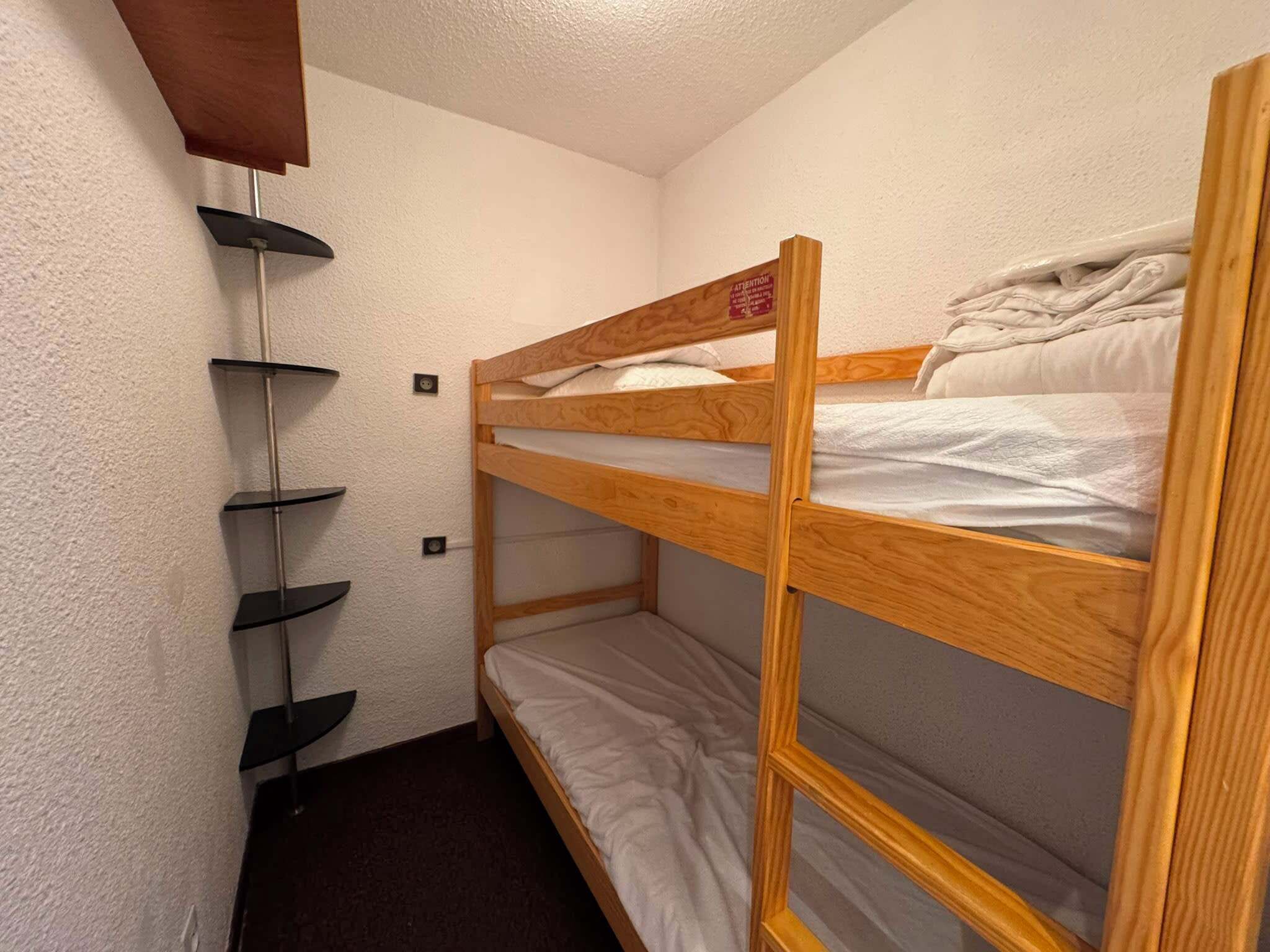 A bunk bed with a wooden frame and white bedding, a black metal shelf unit against the wall, and a small power outlet on the wall.