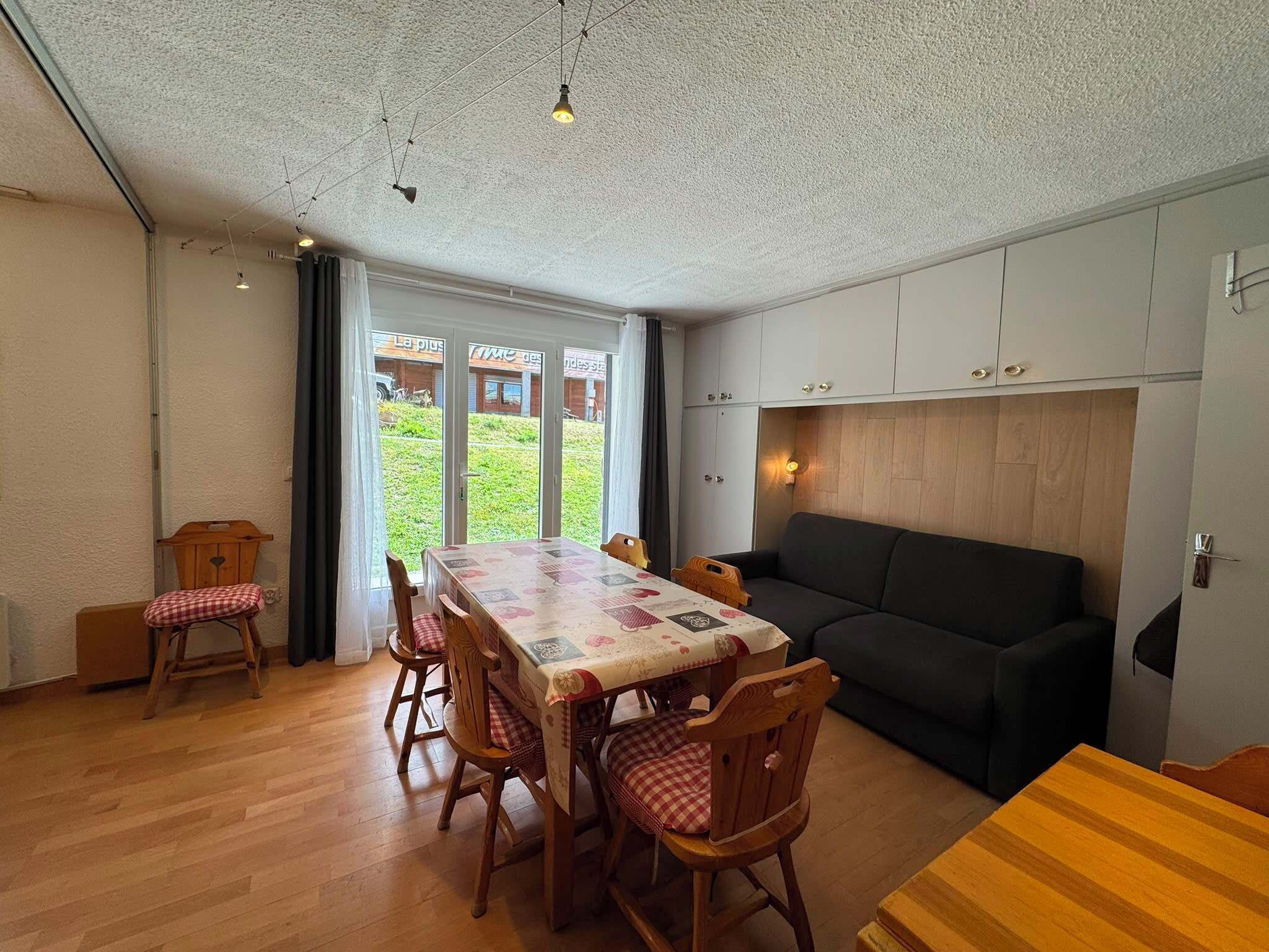 A dining area with a table and chairs, a sofa, and a view of a garden through a glass door.