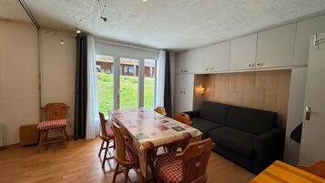 A dining area with a table and chairs, a sofa, and a view of a garden through a glass door.