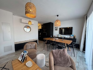 A modern living room with a dining area, featuring a flat-screen TV, wicker pendant lights, and a round mirror on the wall. - Chalet Élégance - 8 pers - 3 chambres - 2 salles de bains (Risoul 1850)