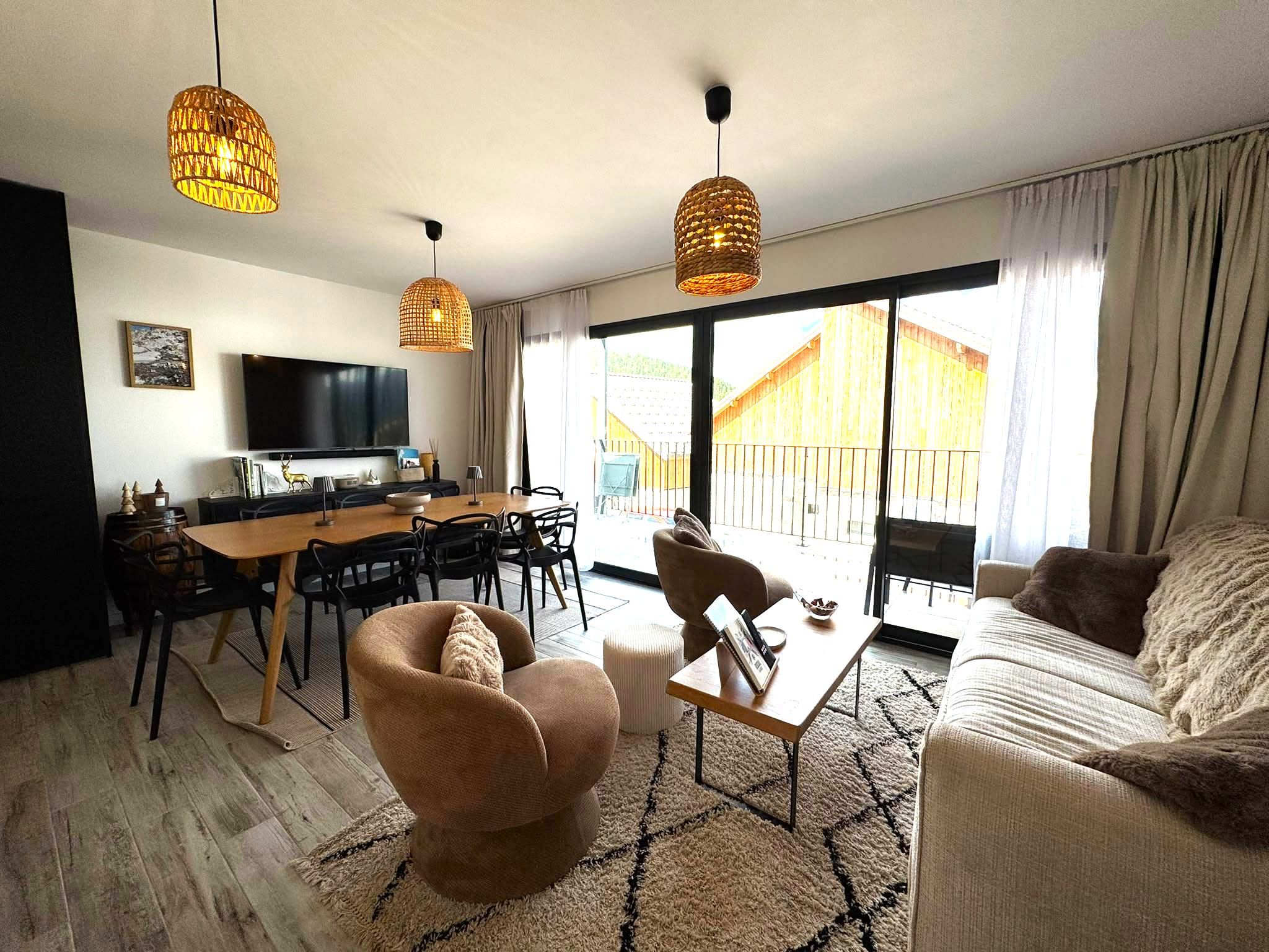 Ein modernes Wohnzimmer mit einem Holzesstisch, gemütlicher Sitzgelegenheit, einem Flachbildfernseher und großen Fenstern mit Blick auf ein Holzgebäude außerhalb.