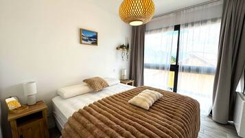 A bedroom with a large bed, a bedside table with a lamp, a hanging plant, and a window with curtains.