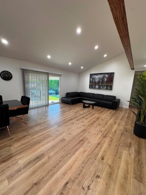 Ein geräumiges Wohnzimmer mit Holzboden, einer schwarzen Couch, einem Esstisch mit Stühlen, einem Bild an der Wand und einem großen Fenster mit Jalousien.