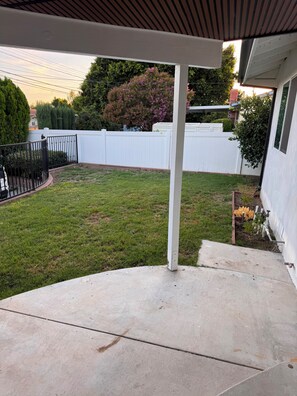 Ein überdachter Patio-Bereich mit Betonboden, weißem Zaun und einem Baum mit rosa Blüten.