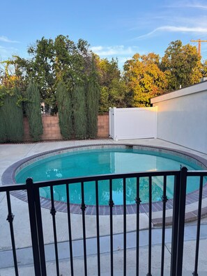 Ein Swimmingpool mit schwarzem Metallzaun im Vordergrund, umgeben von einer Ziegelmauer und Bäumen.
