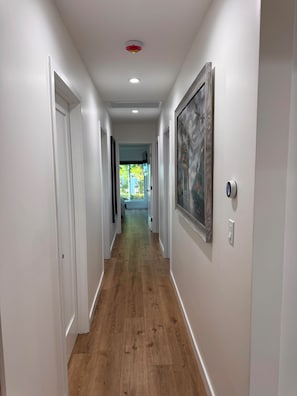 A hallway with white walls, wooden flooring, and a door at the end leading to a bright room with a view of greenery. - Awesome 4-bedroom house with AC in charming Los Angeles (Los Angeles)