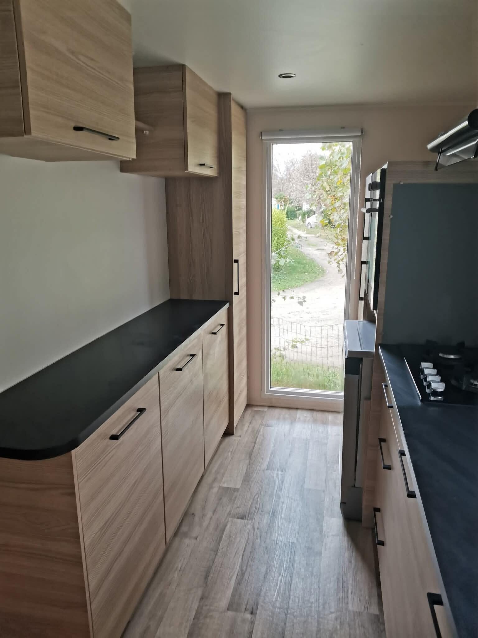 Une cuisine moderne avec des meubles de couleur bois clair, un plan de travail noir et une vue sur un jardin grâce à une porte vitrée.